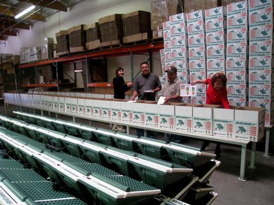 People Standing In A Warehouse Surrounded By Massager Boxes